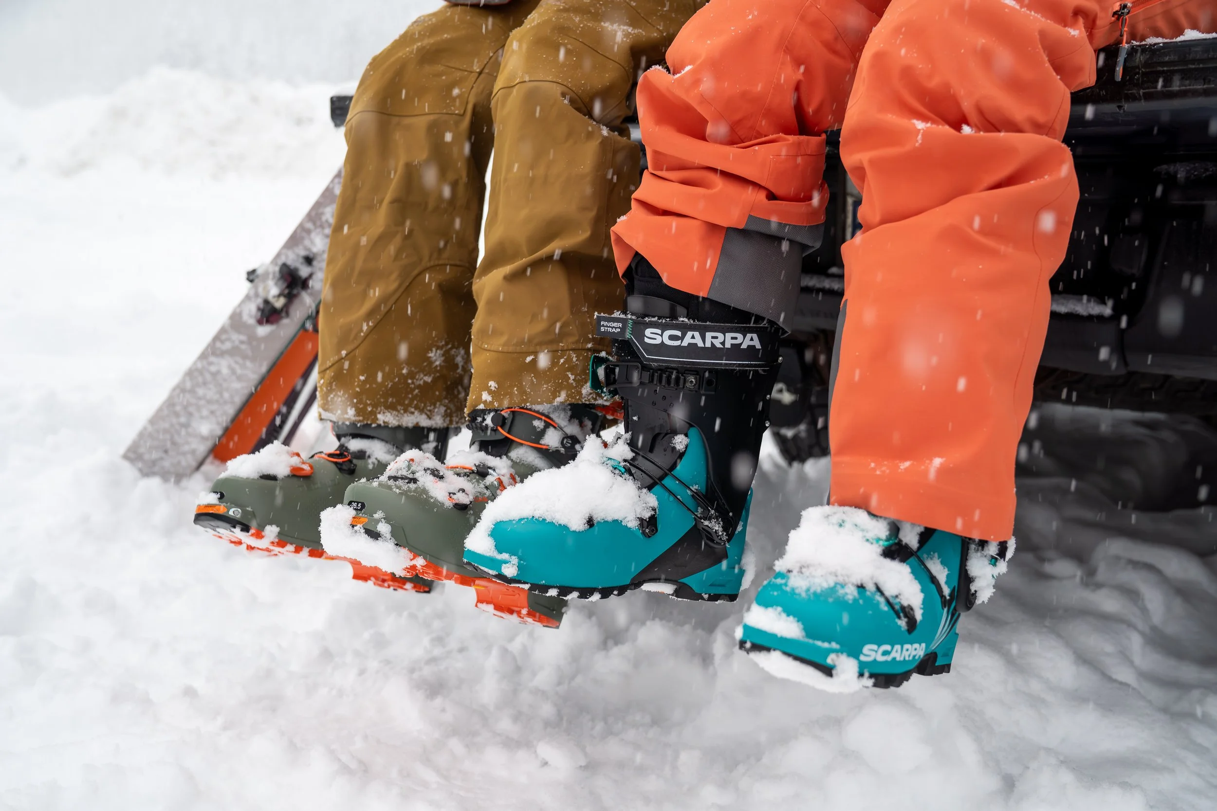 A photo of two people in ski clothes and ski boots sitting on the tailgate of a truck. The ski boots and parking lot are covered in snow.