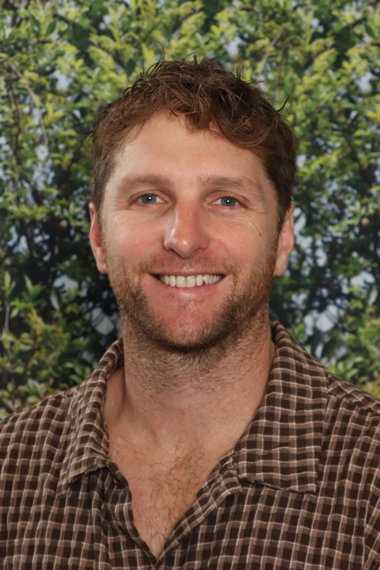 A smiling man with short brown hair and a beard, wearing a brown checkered shirt, standing outdoors with green foliage in the background.