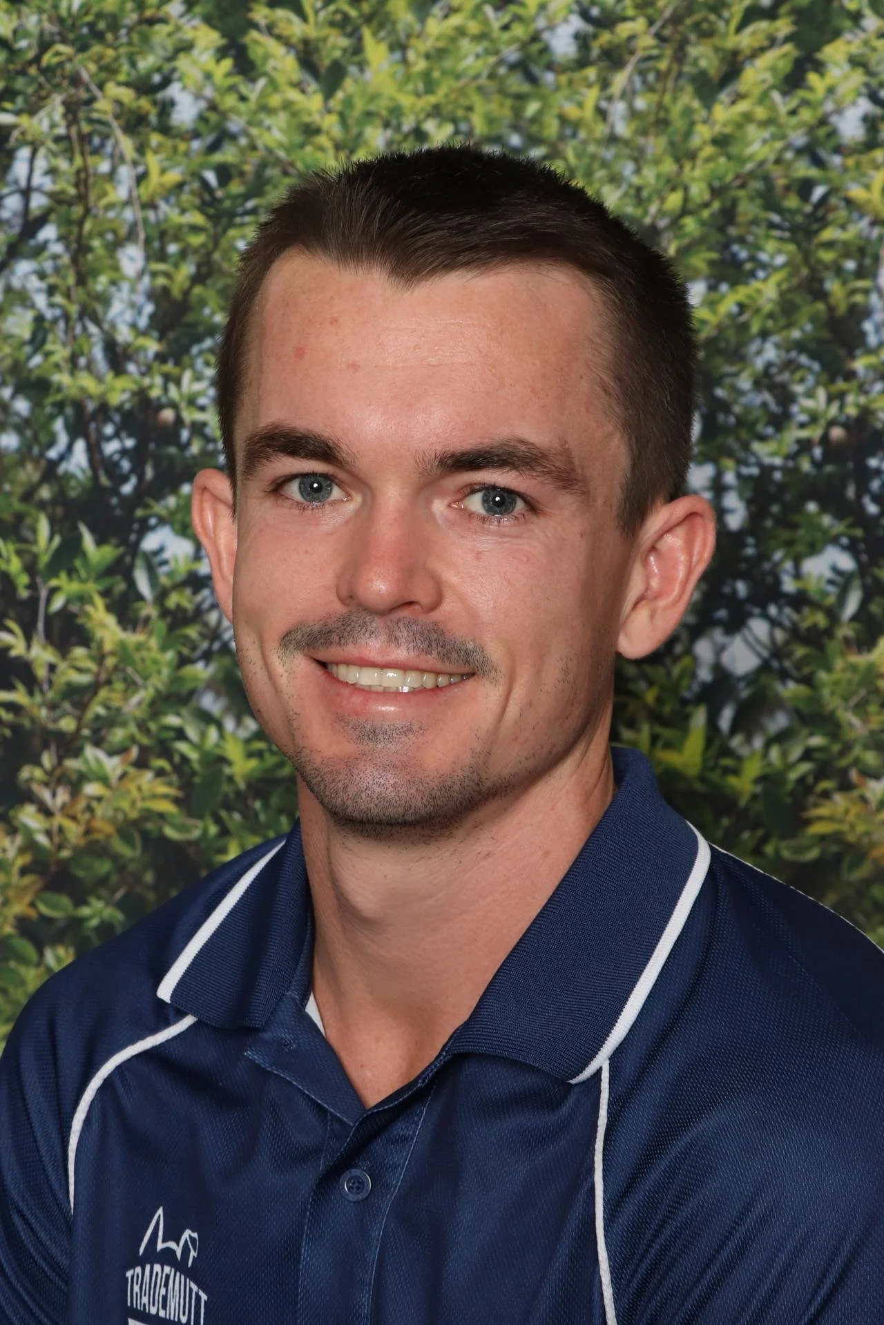 A man with short dark hair, blue eyes, and light skin, smiling, wearing a navy blue shirt with white piping and a logo that says 'TradeMutt' in front of a leafy green background.