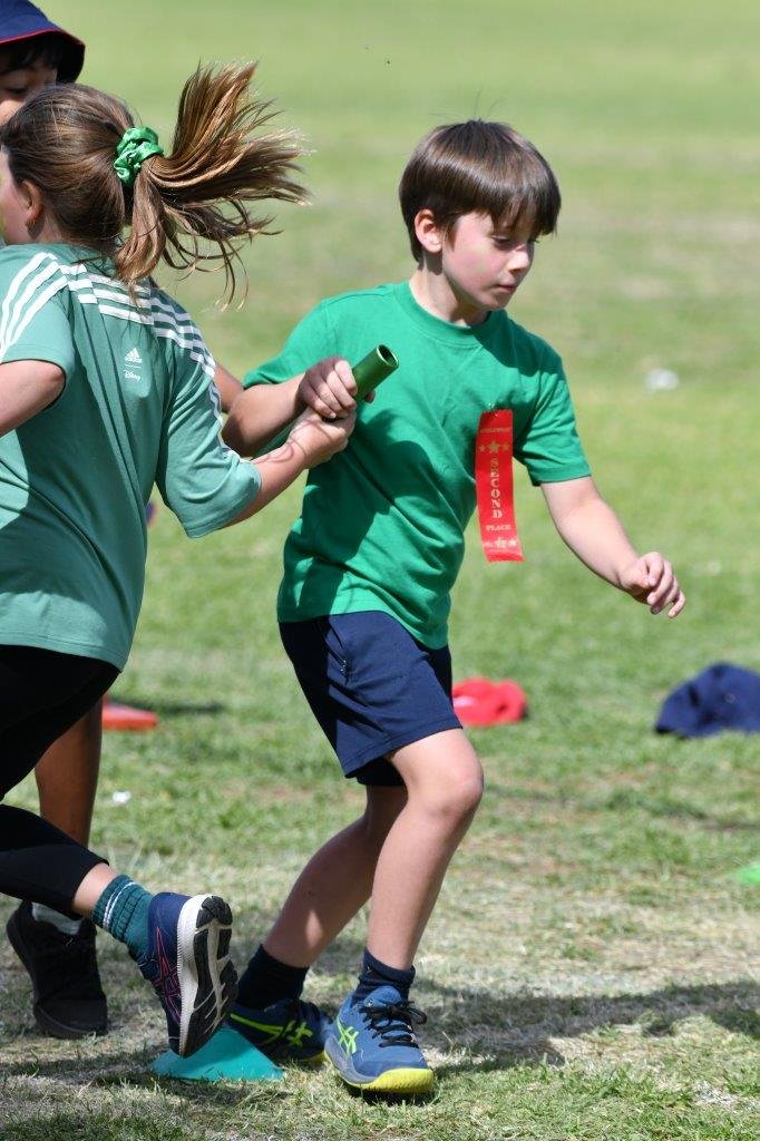 Children participating in an outdoor relay race, with one boy in a green shirt and red ribbon running, and a girl in athletic clothing about to pass a baton.