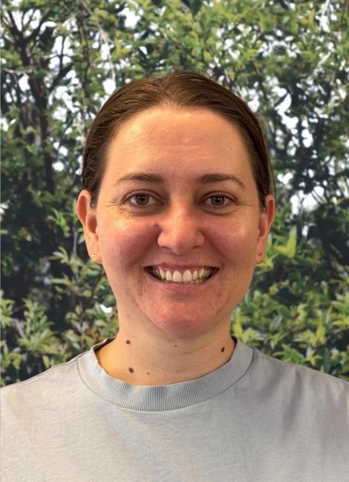 Smiling woman with brown hair and light skin, wearing a gray shirt, standing in front of a green, leafy tree background.