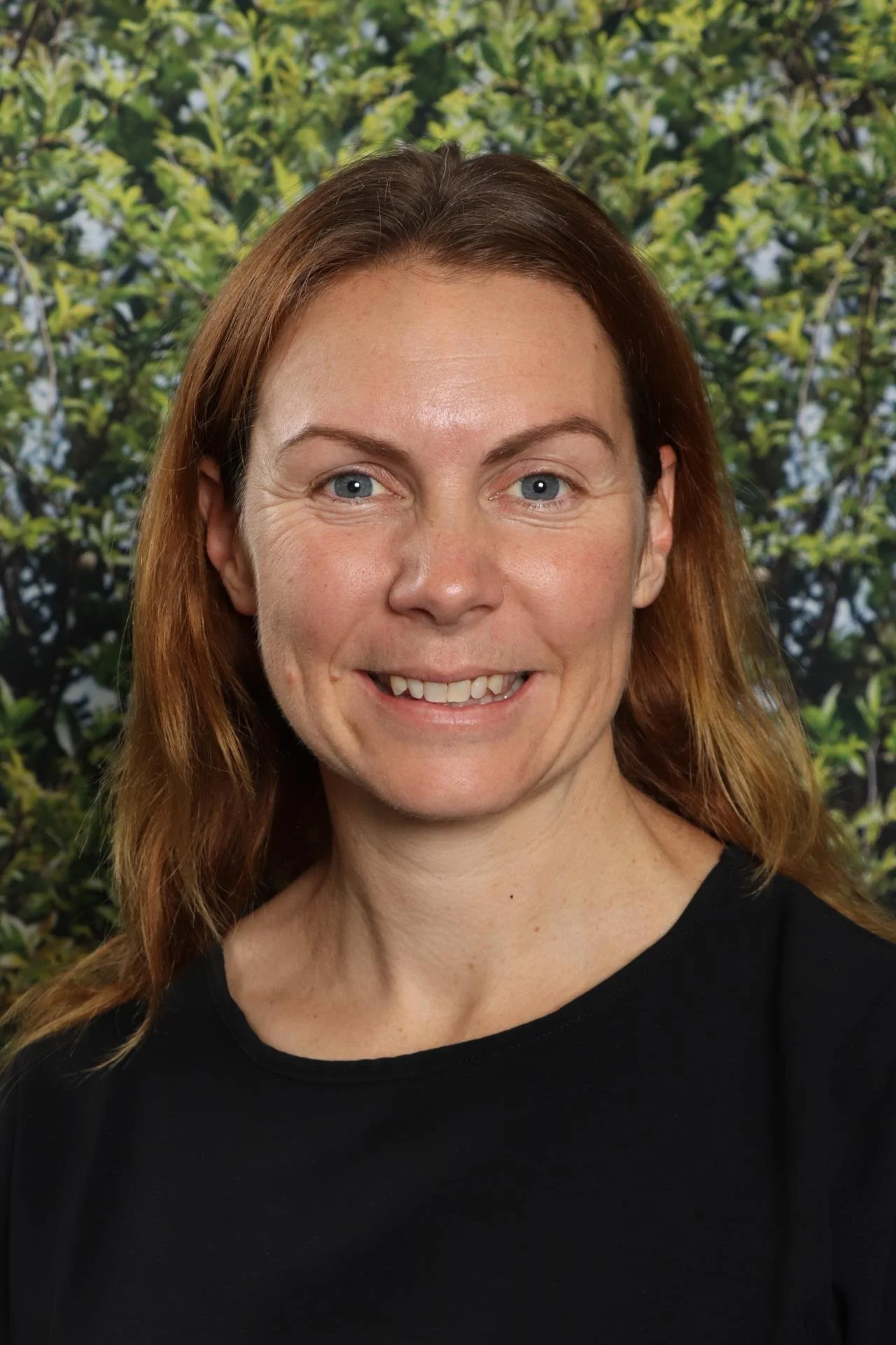 A smiling woman with red hair and blue eyes, wearing a black top, standing in front of a background of green foliage.