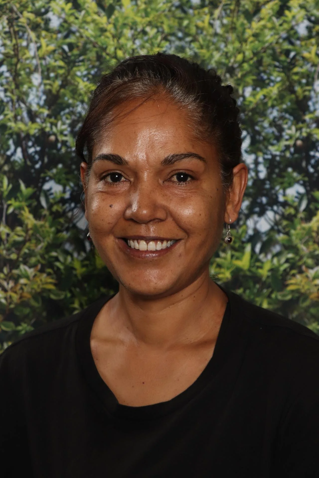 A woman with short dark hair, wearing earrings and a black top, smiling in front of a leafy green background.