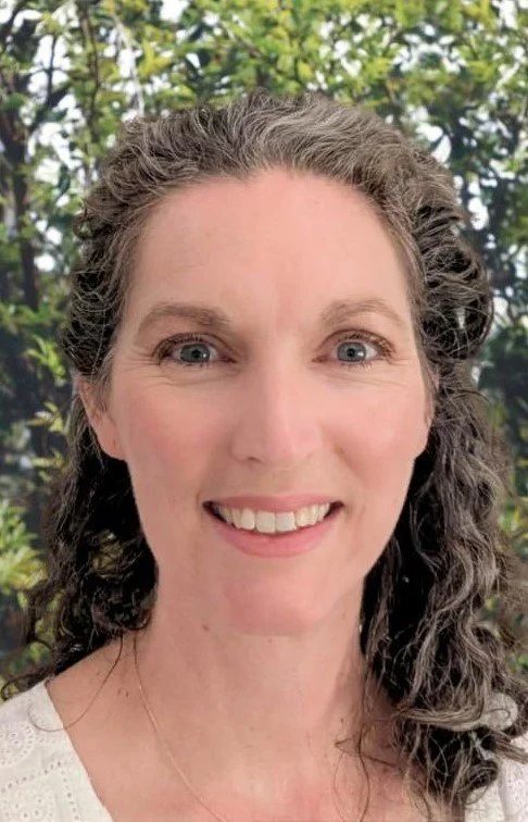 A woman with gray curly hair smiling outdoors with green foliage in the background.