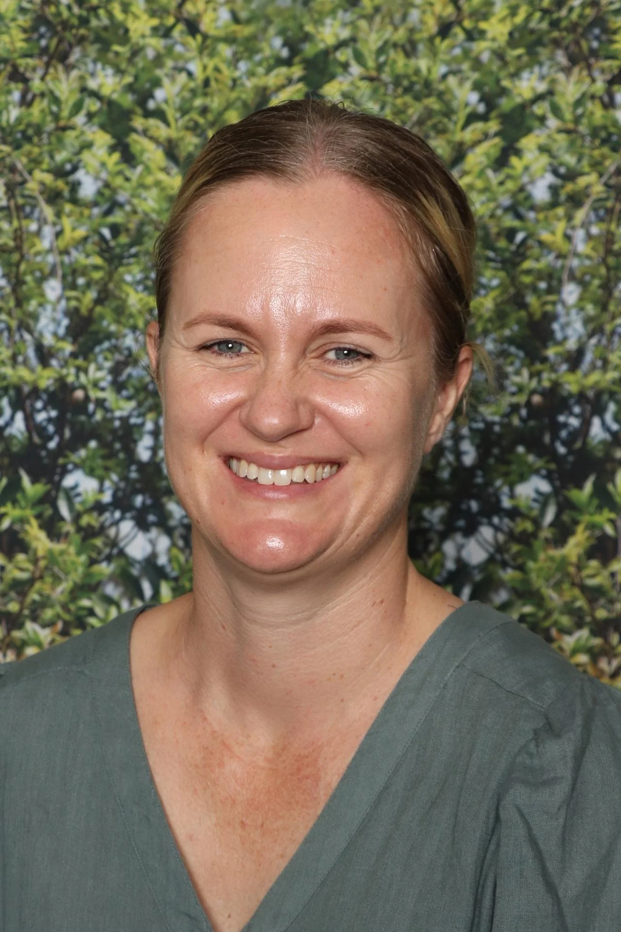 Close-up of a smiling woman with light skin and brown hair, standing outdoors with blurred green foliage in the background.
