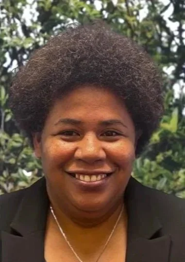 A woman smiling outdoors with green foliage in the background, wearing a black blazer and a necklace.