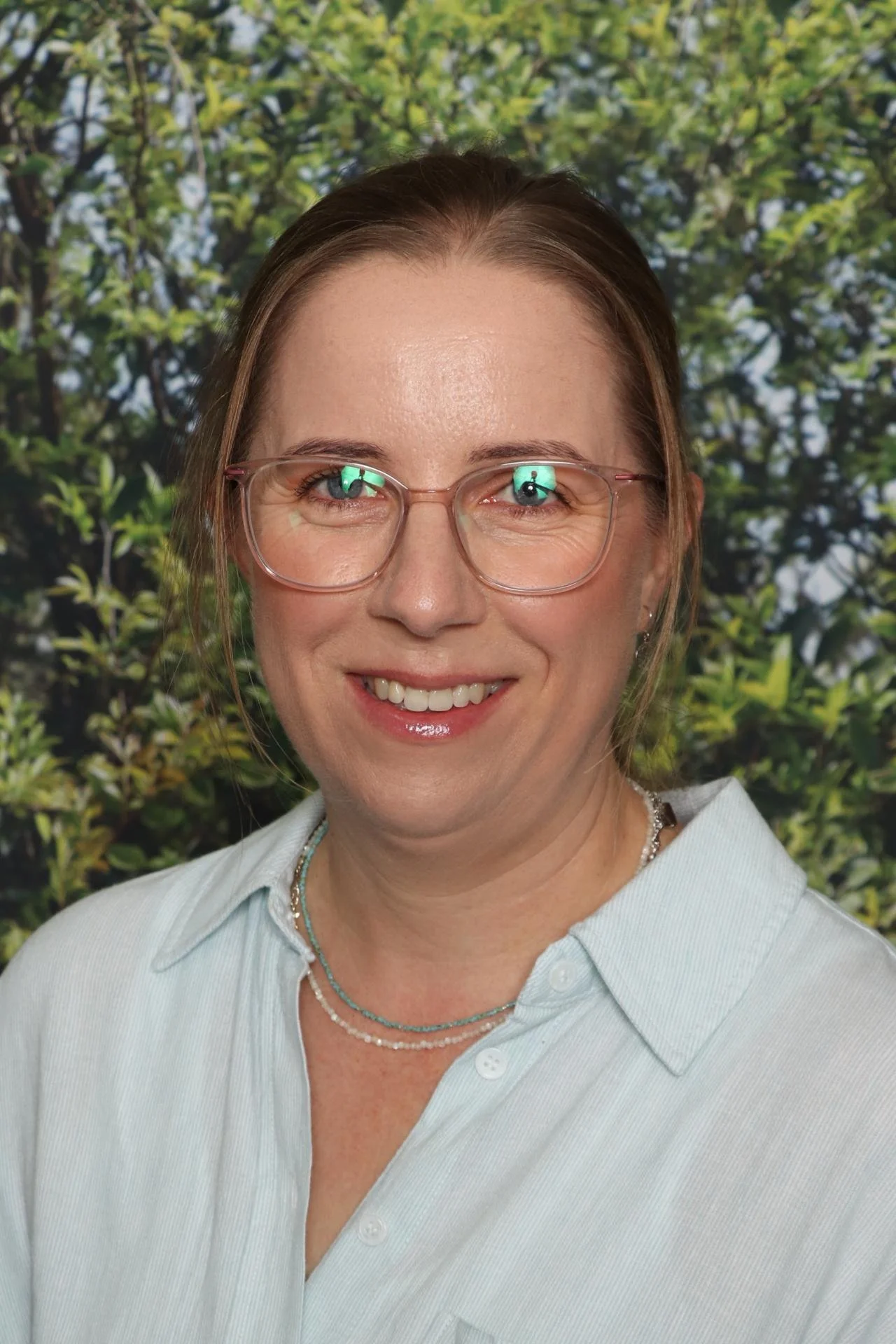 A smiling woman with light brown hair, wearing pink glasses, a light blue collared shirt, and layered necklaces, standing in front of a background of green foliage.