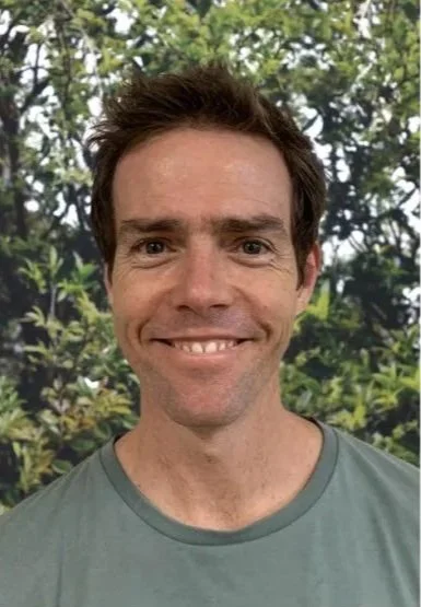 A man with brown hair smiling outdoors with green trees in the background.
