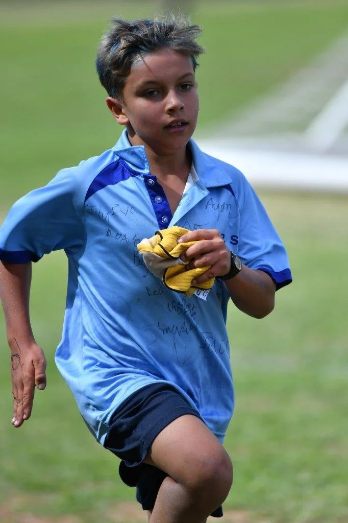 Young boy running outdoors on a grassy field, wearing a blue sports shirt and black shorts, holding yellow gloves, with short messy brown hair.