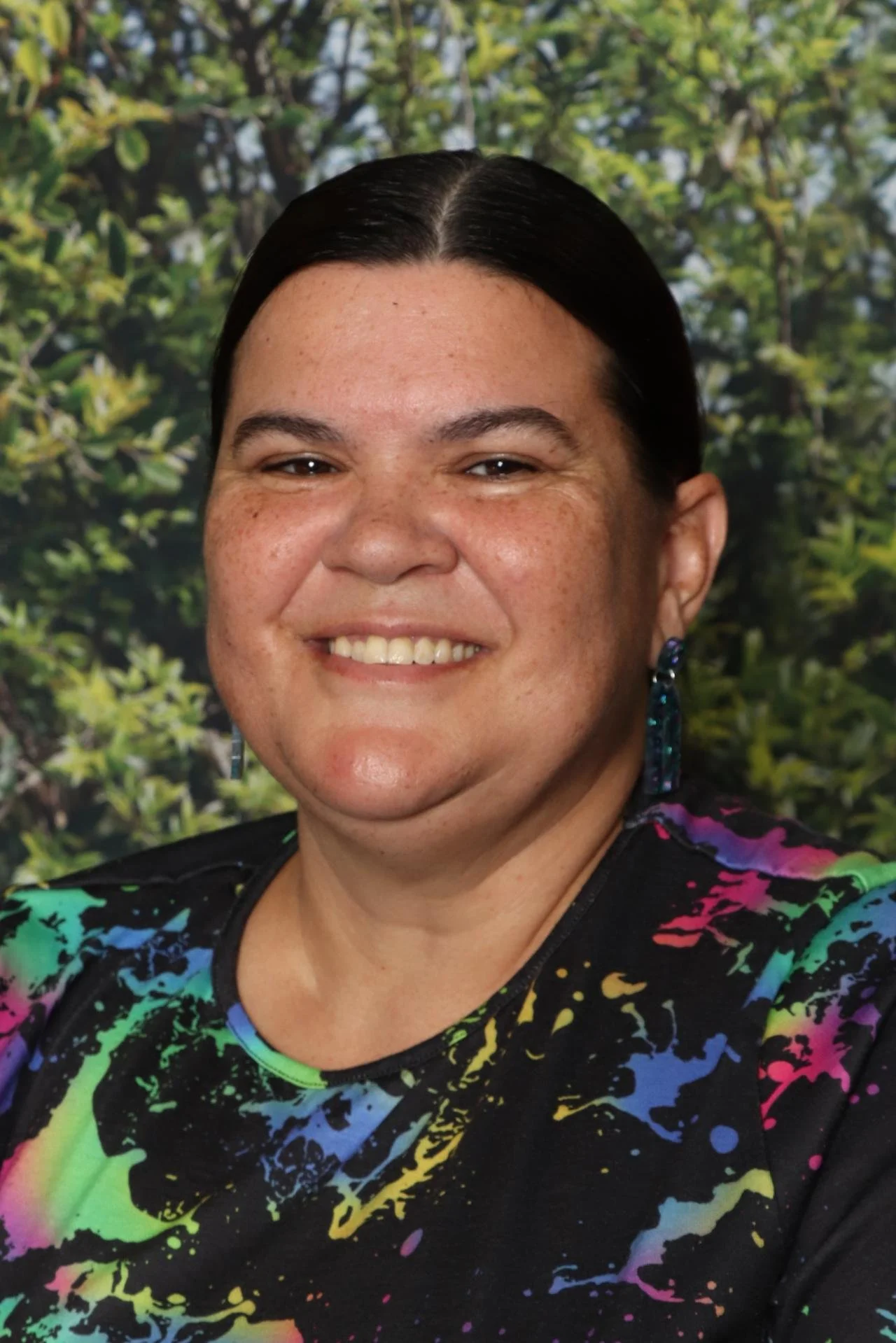 A woman with dark hair and earrings smiling in front of a green leafy background, wearing a colorful, splatter-patterned top.