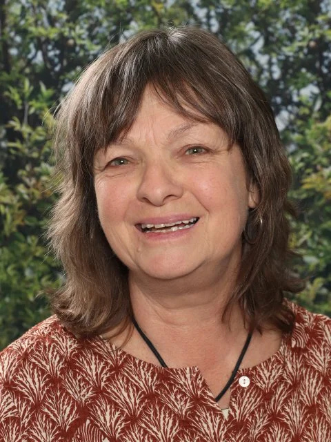 A smiling middle-aged woman with shoulder-length brown hair and light skin, wearing a patterned blouse and a black necklace, standing outdoors with green foliage background.