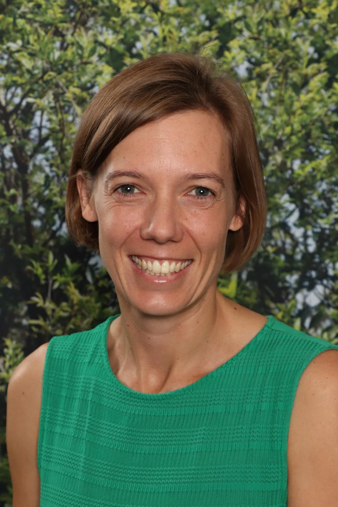 Close-up of smiling woman with short brown hair wearing a green sleeveless top, standing outdoors with green trees in the background.