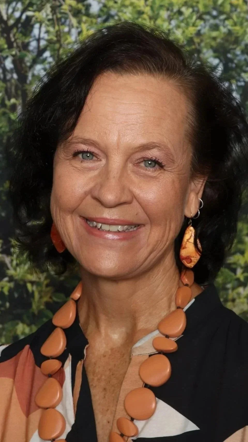 Close-up photo of a smiling woman with black hair, wearing orange jewelry, and a multicolored top, with a background of green trees.
