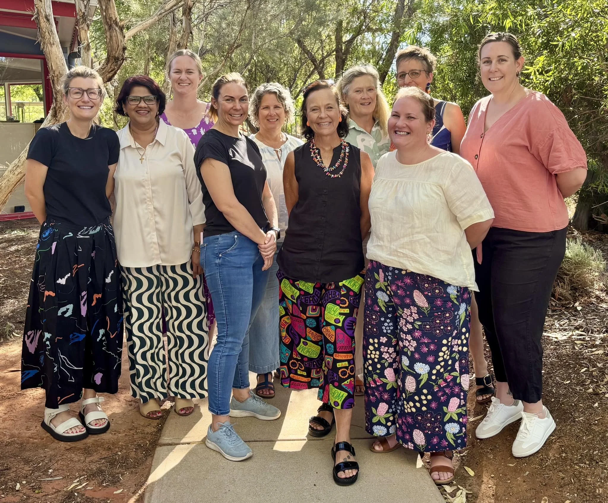 A group of eleven women smiling outdoors in a lush, green setting with trees. They are dressed casually and standing close together.