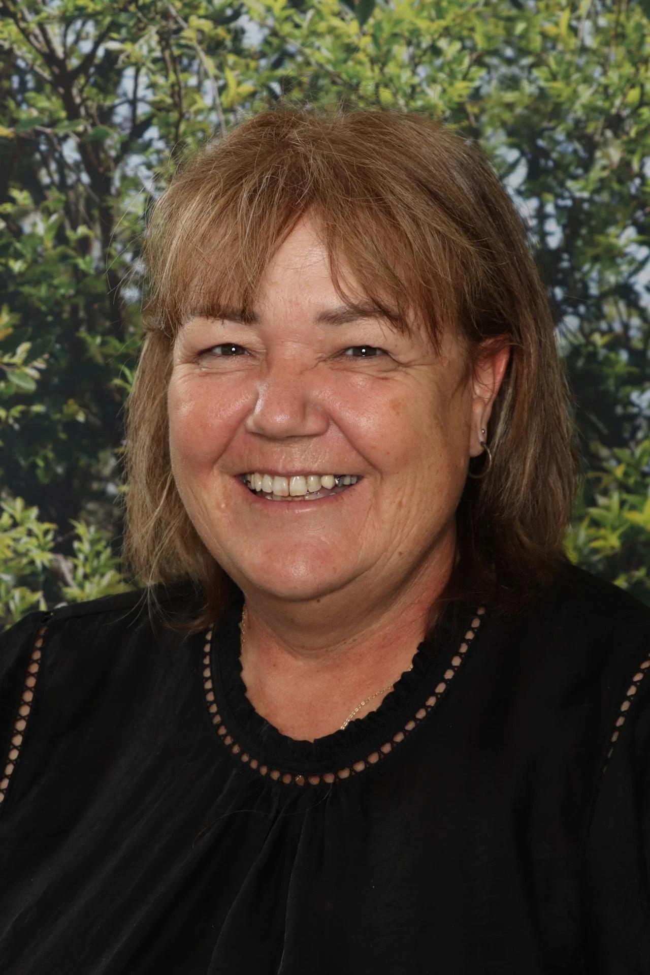 A smiling woman with shoulder-length reddish-brown hair, wearing a black top with lace details, stands in front of a green leafy background.