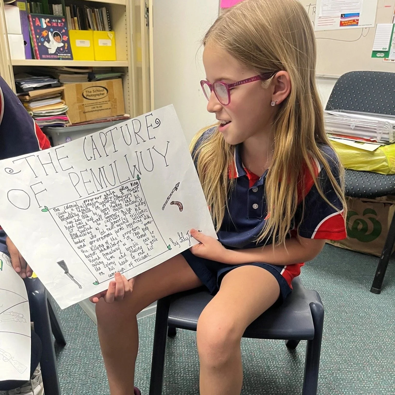 Girl in glasses sitting on a chair, holding a large poster board titled 'The Capture of Penuluky,' inside a classroom with shelves, books, and files around her.