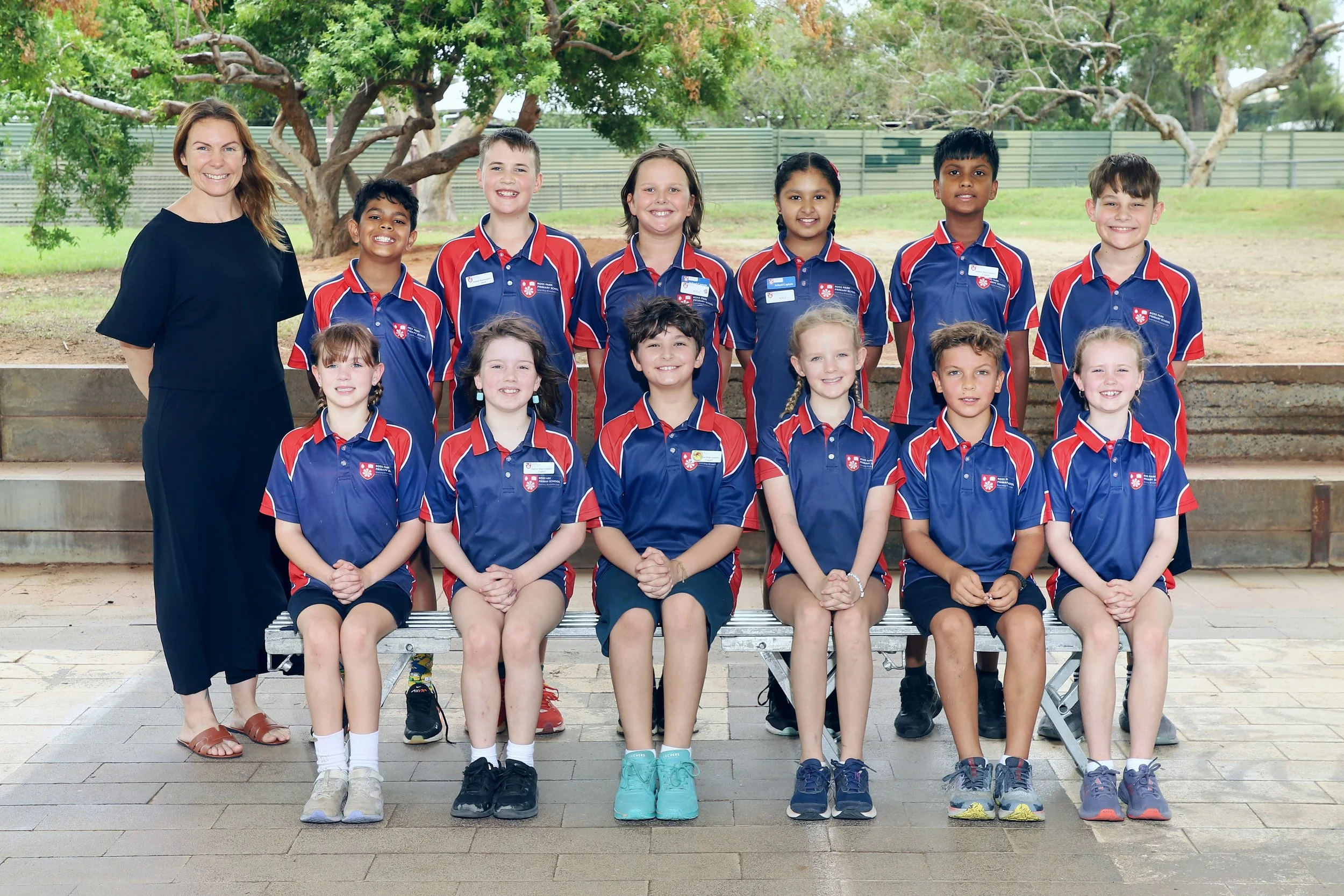 A group of young school children and their teacher posing for a class photo outdoors, in front of trees and a fence, all wearing school uniforms in blue and red.