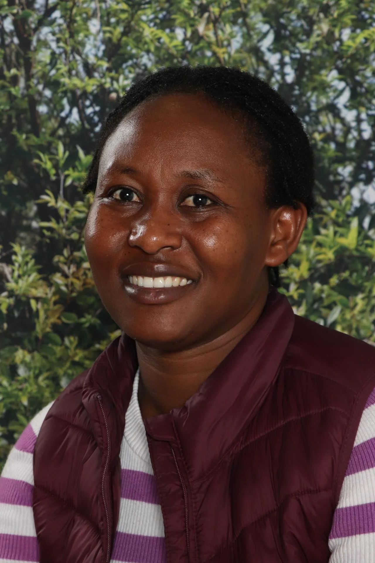 Close-up of a smiling woman with short, dark hair, wearing a maroon vest over a white and purple striped sweater, standing outdoors with green leafy trees in the background.