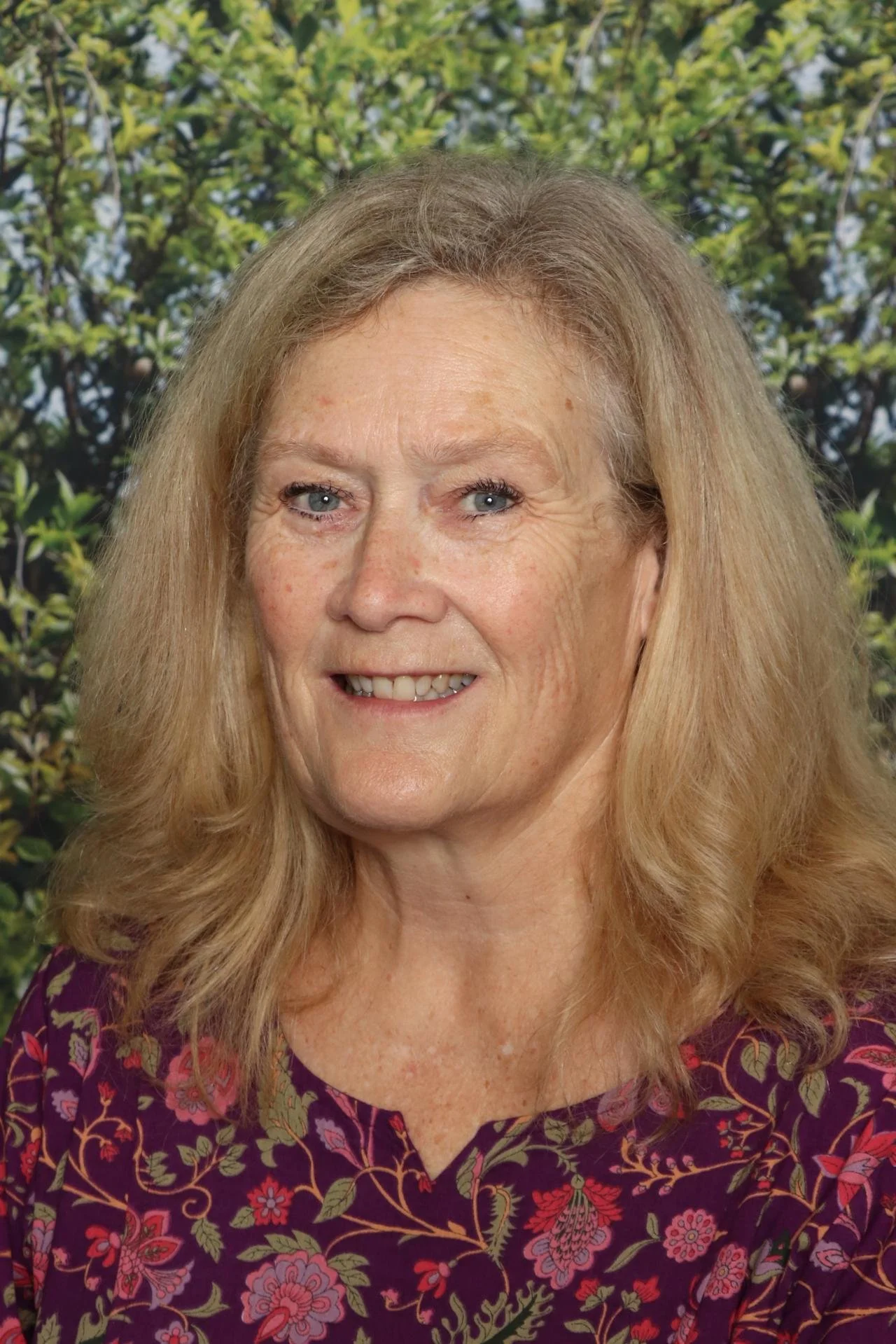 A portrait of an older woman with long blonde hair, blue eyes, and a warm smile, wearing a purple floral top, standing in front of a background of green foliage.