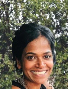 A woman with dark hair styled in an updo, smiling outdoors with green trees in the background.