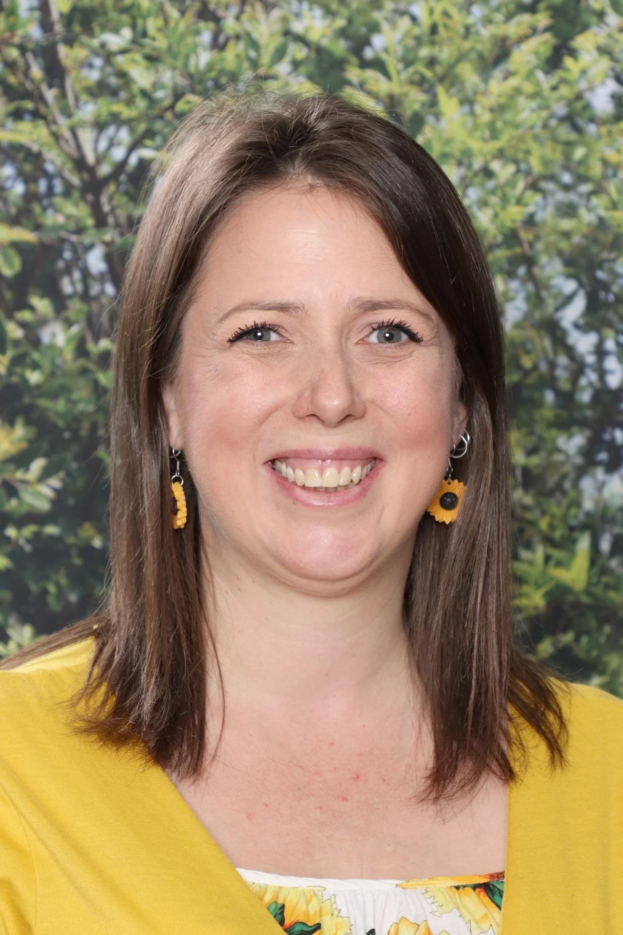 A woman with long brown hair, blue eyes, and earrings with sunflower designs, smiling in front of a leafy green background.