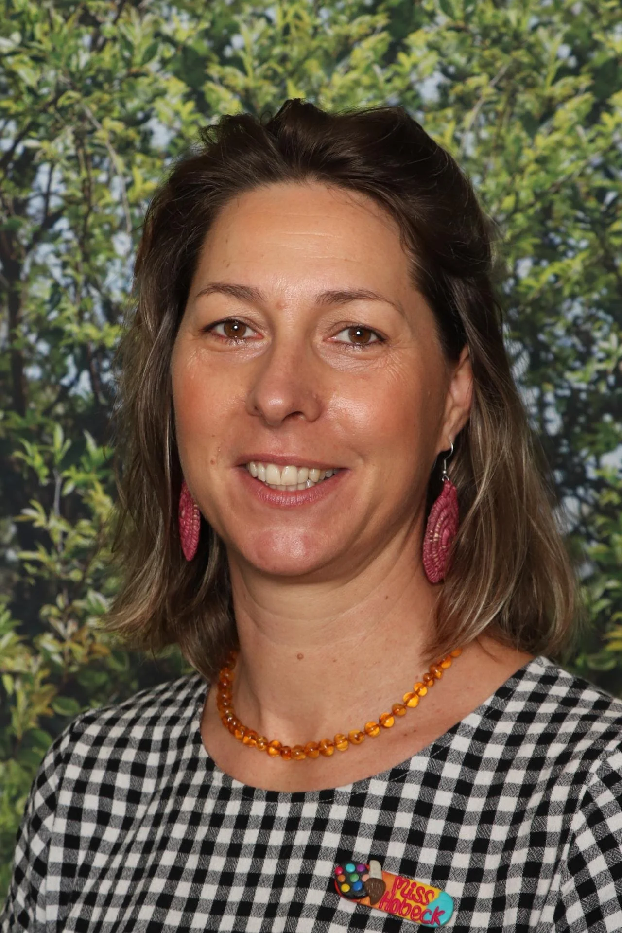 A woman with brown hair, wearing pink earrings and an orange bead necklace, smiling at the camera. She is dressed in a black and white checkered top with a colorful badge that reads 'Miss Hoops' and features images of candies. The background has green leaves.
