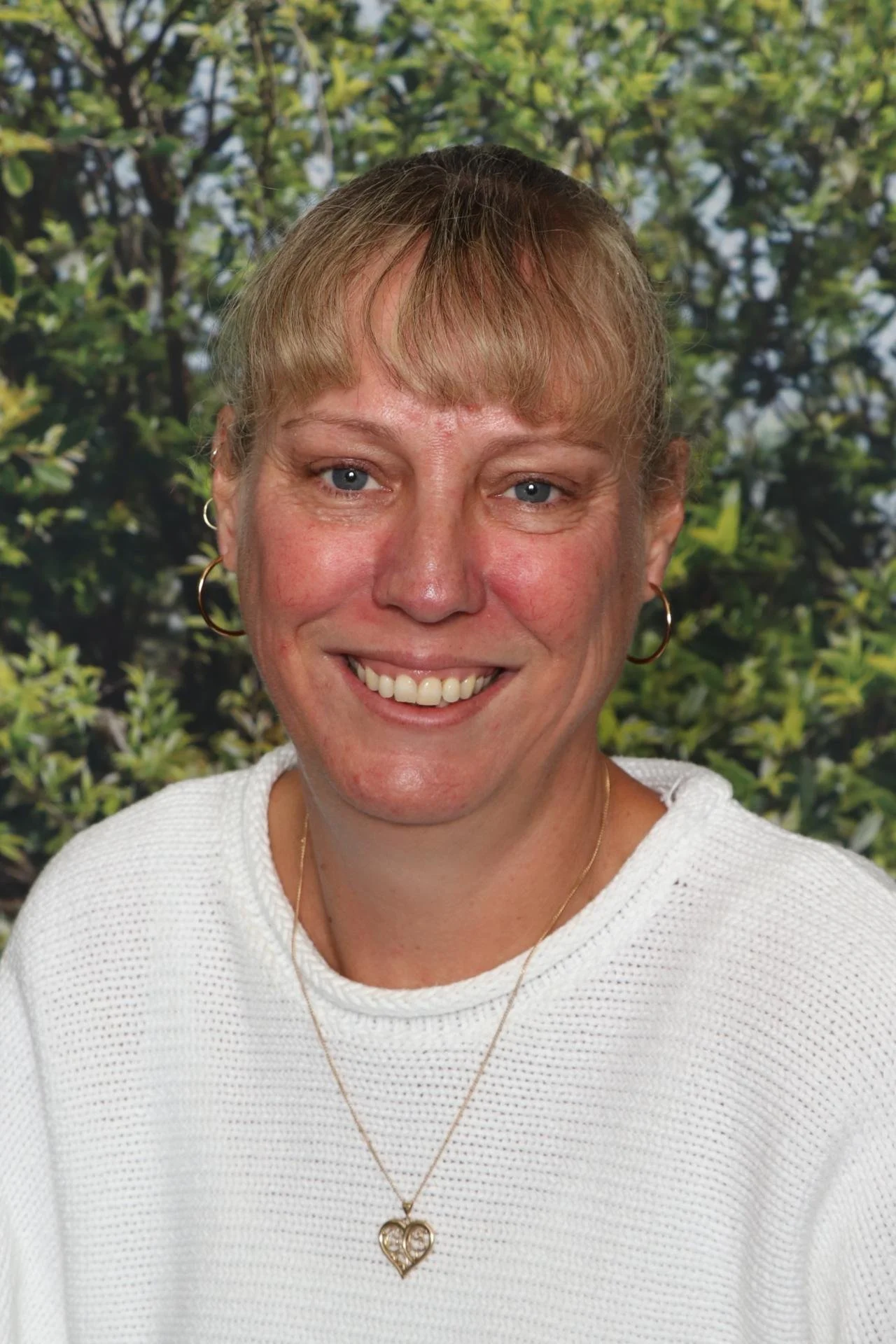 A woman smiling with light brown hair and blue eyes, wearing gold hoop earrings, a gold necklace with a heart-shaped pendant, and a white textured sweater, standing outdoors with green foliage in the background.