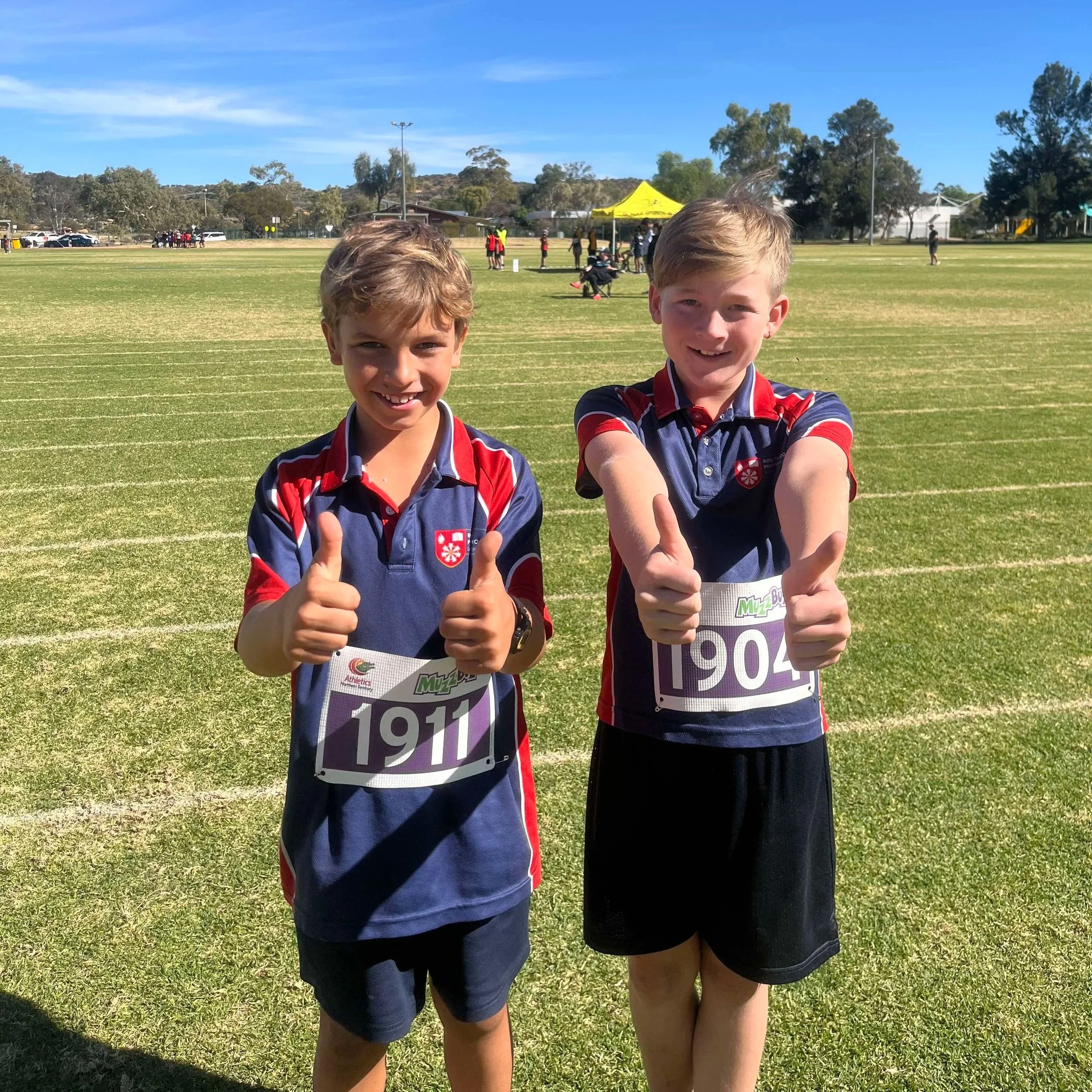 Two young boys in athletic uniforms taking a selfie together on a grassy sports field, with other people and a yellow tent visible in the background.