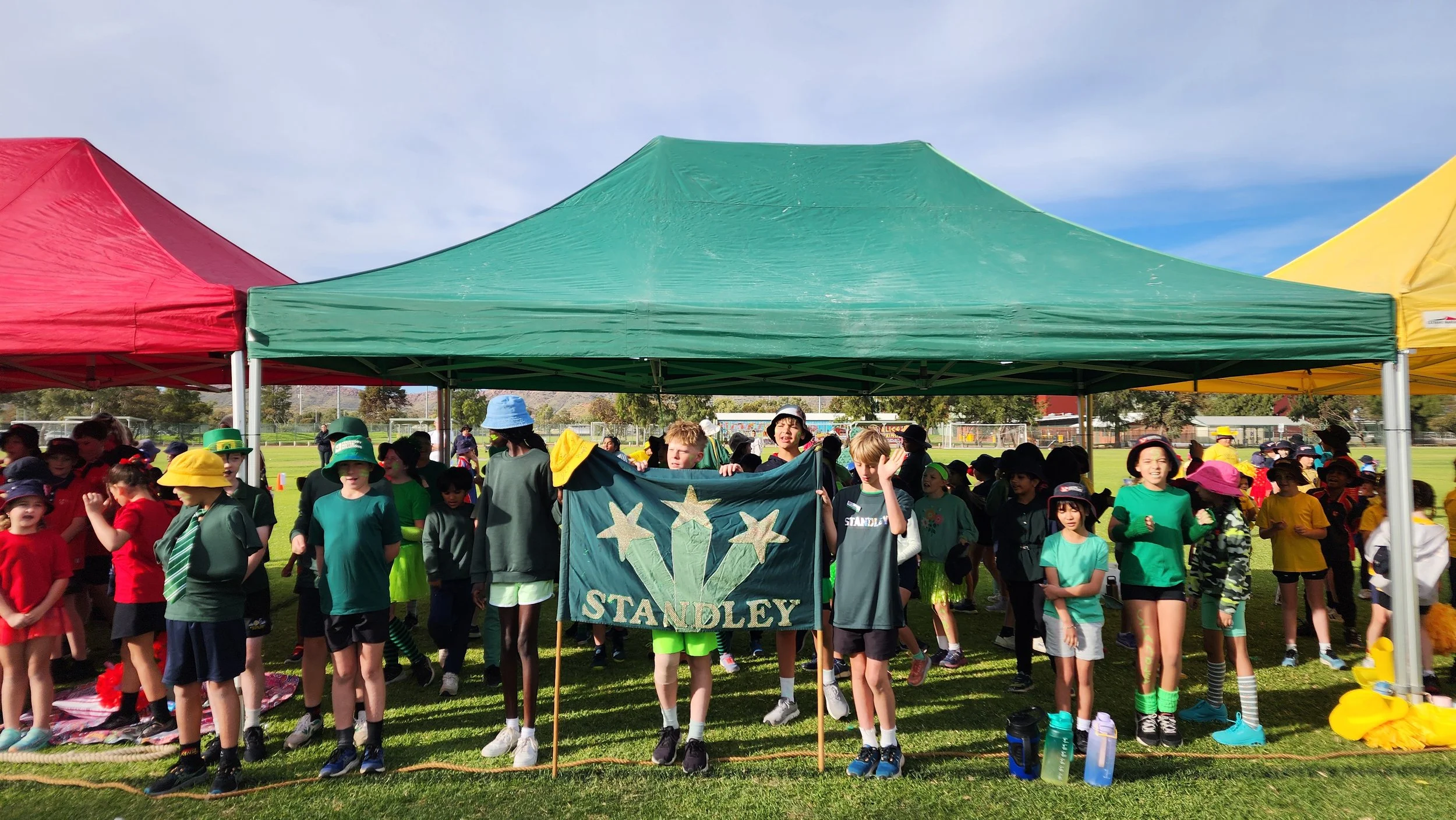 A group of children gathered under a green canopy at an outdoor event, holding a banner that says 'Stanley.' Many children are wearing colorful hats and casual clothing, on a grassy field with a sports field and trees in the background.