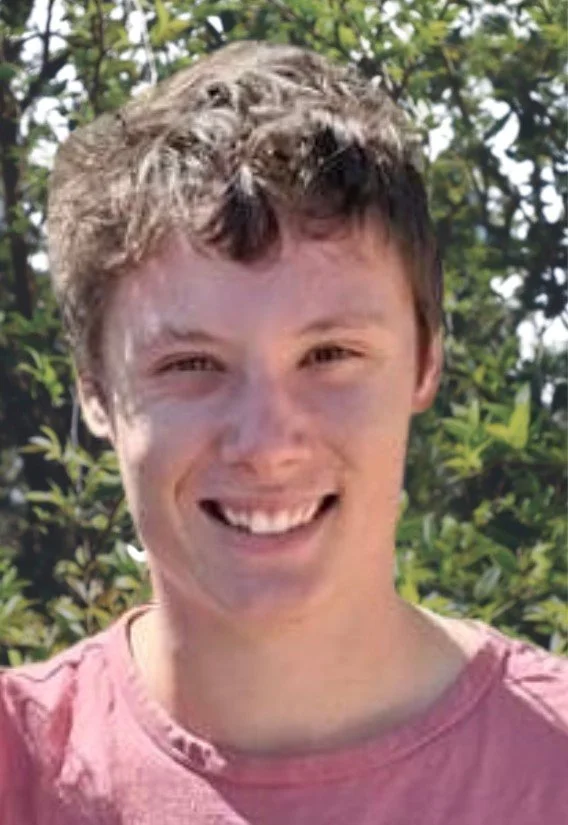 Young man with short, curly brown hair smiling outdoors with green trees in background.