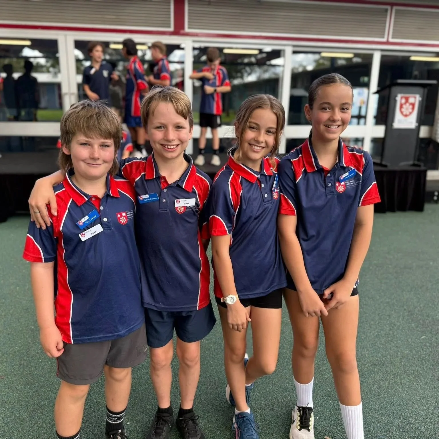 Four children in an outdoor setting wearing sports uniforms, smiling and standing side by side with arms around each other. They are dressed in navy blue and red sports outfits, with two boys and two girls. The background shows windows and other children in similar uniforms.