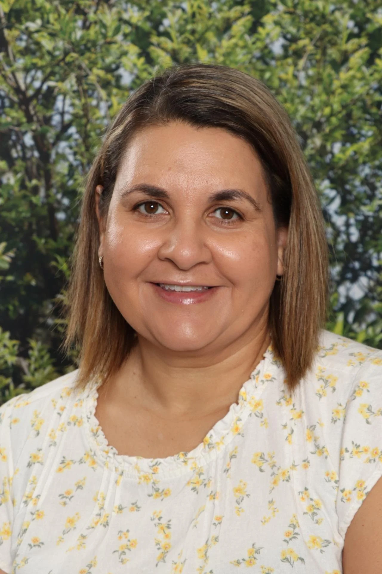 A smiling woman with brown hair and brown eyes wearing a white floral top, standing outdoors with green trees and sunlight in the background.