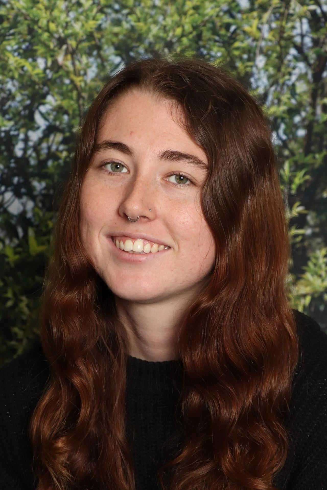 Young woman with long wavy brown hair, green eyes, and a septum piercing, smiling in front of a blurred green foliage background.