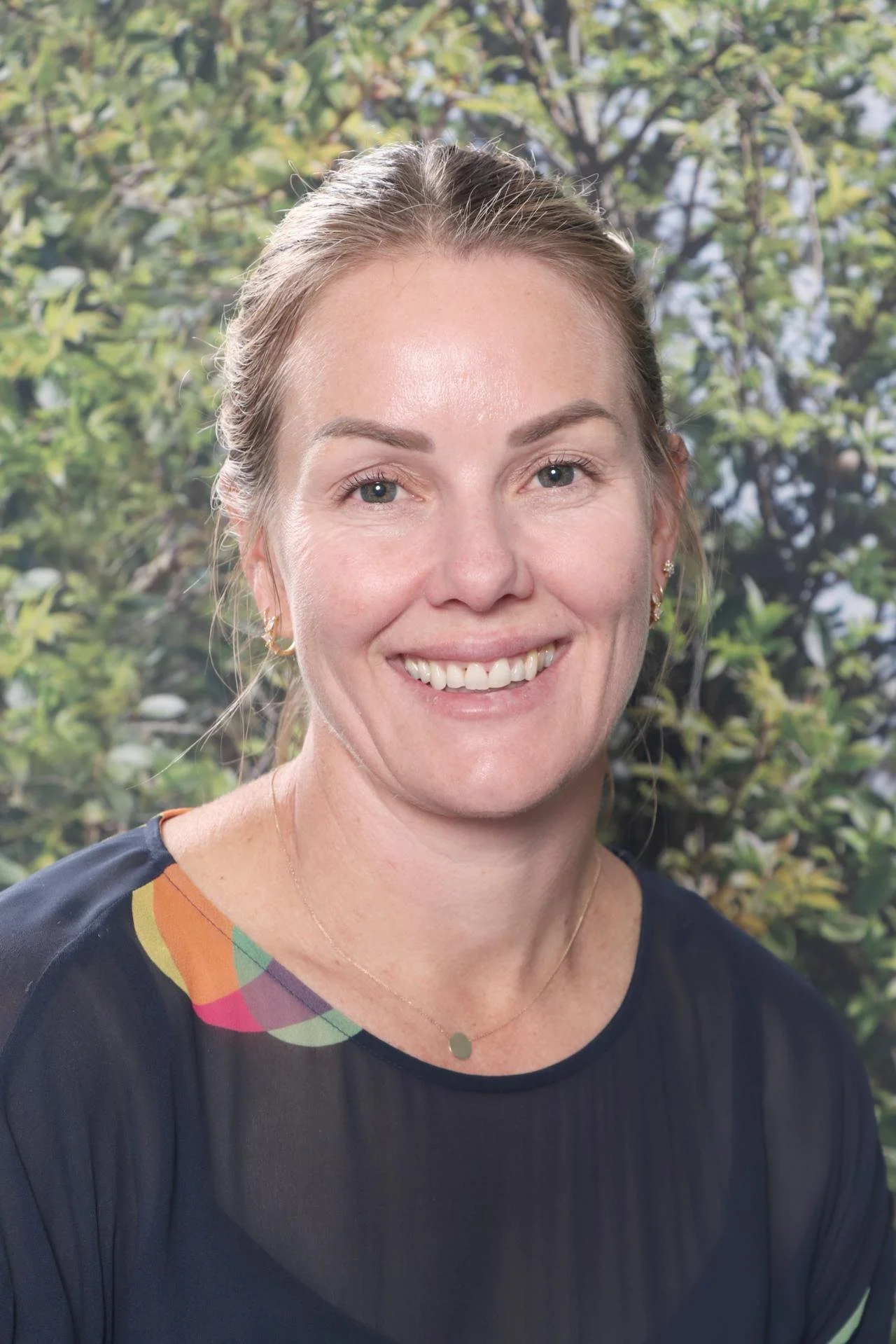 A woman with blonde hair pulled back, smiling, wearing earrings, a delicate necklace, and a black top with colorful accents on the shoulder, standing outdoors with green foliage in the background.