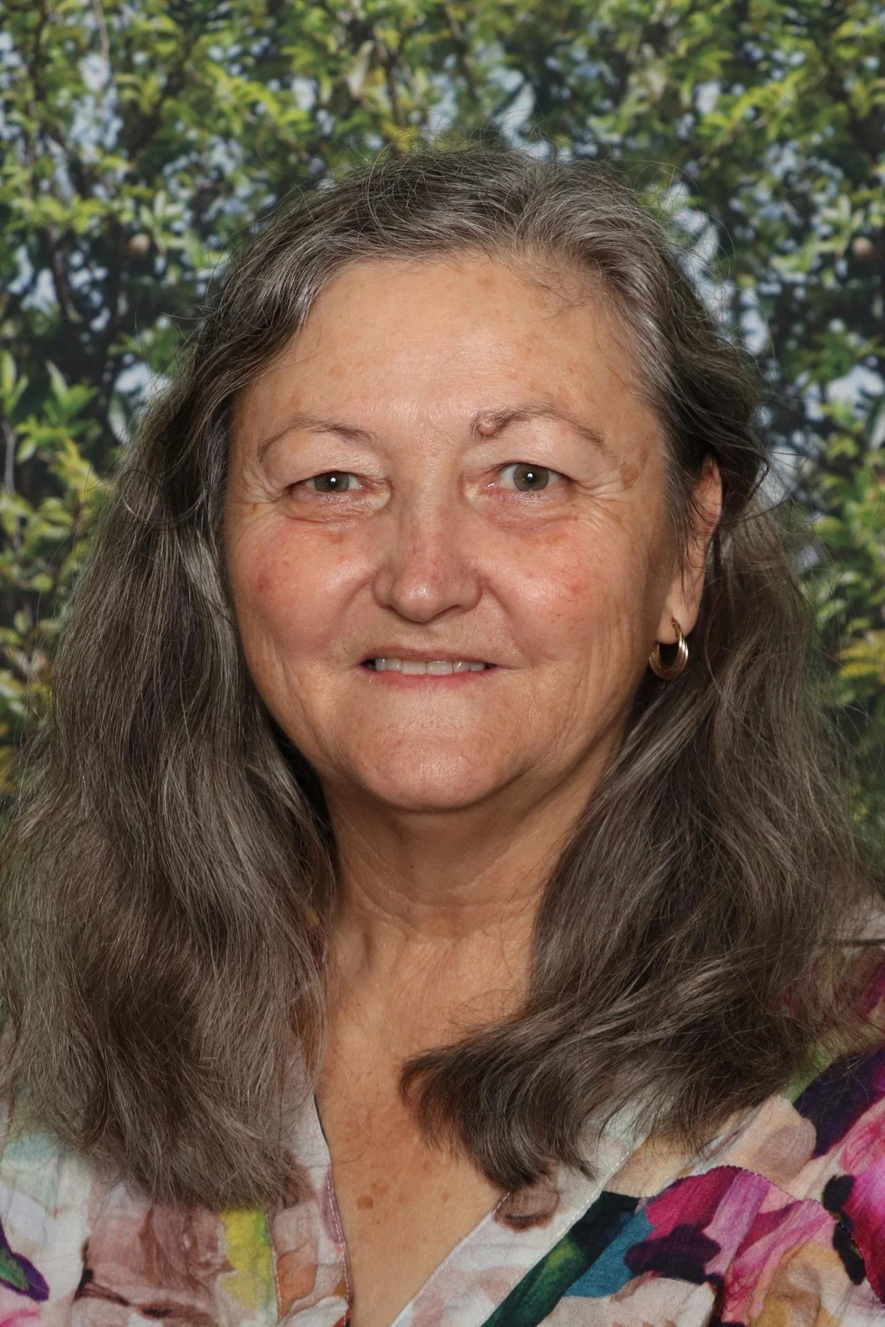 Close-up of a middle-aged woman with long, wavy gray hair, smiling, in front of a background of green foliage.