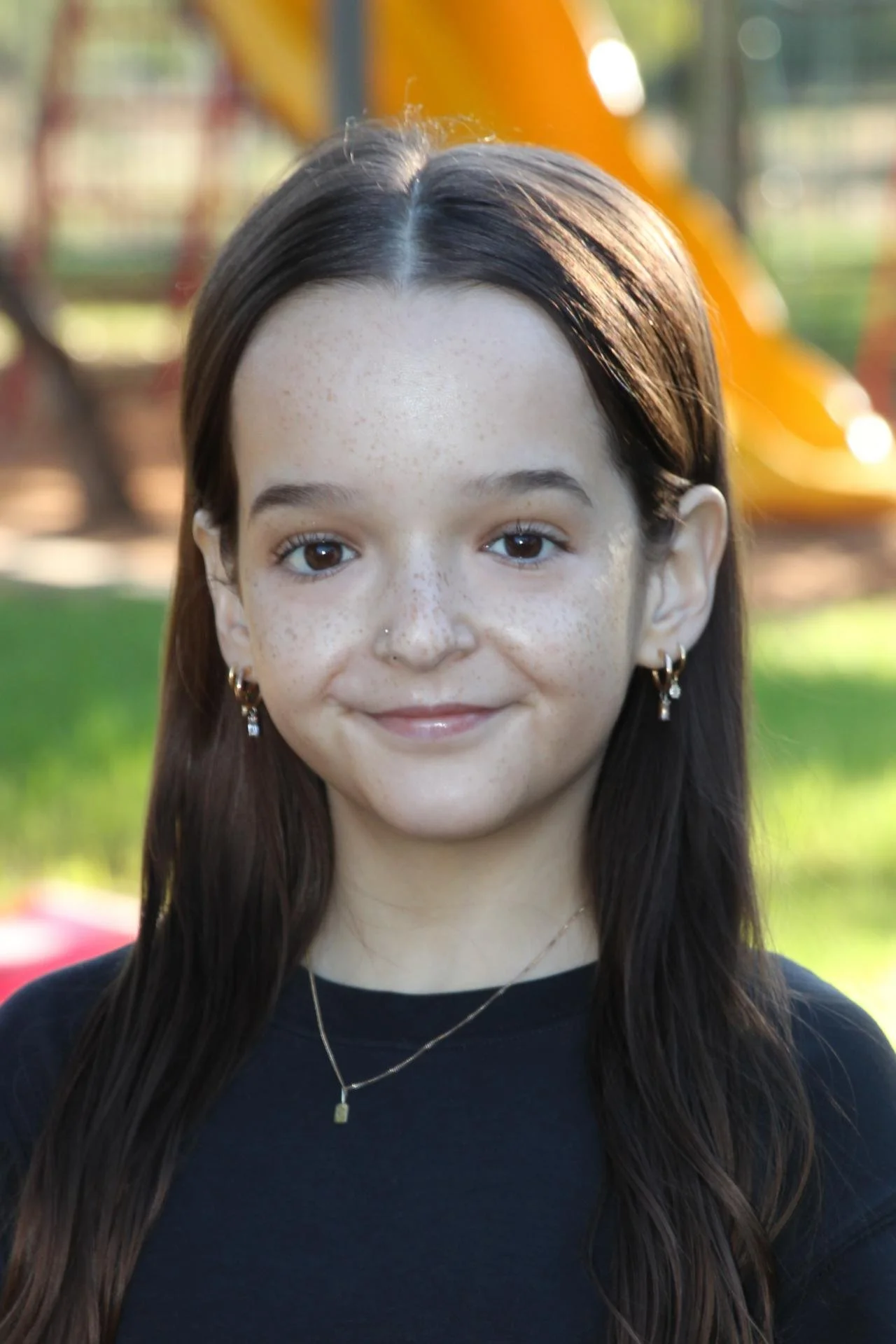 A young woman with long brown hair and freckles, smiling outdoors in front of a playground with a slide.