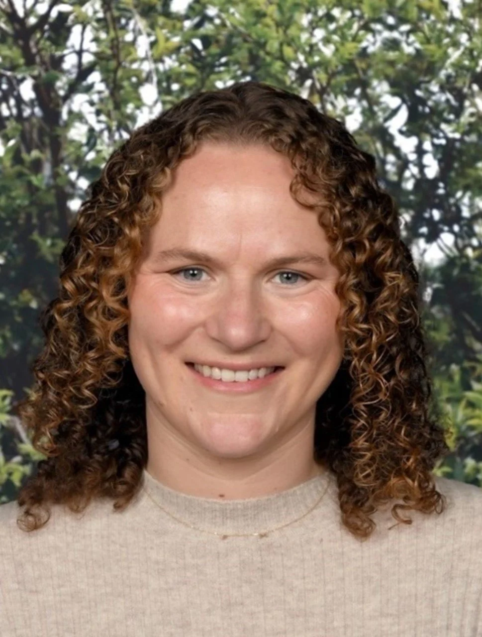 A woman with curly hair smiling in front of a green leafy background.