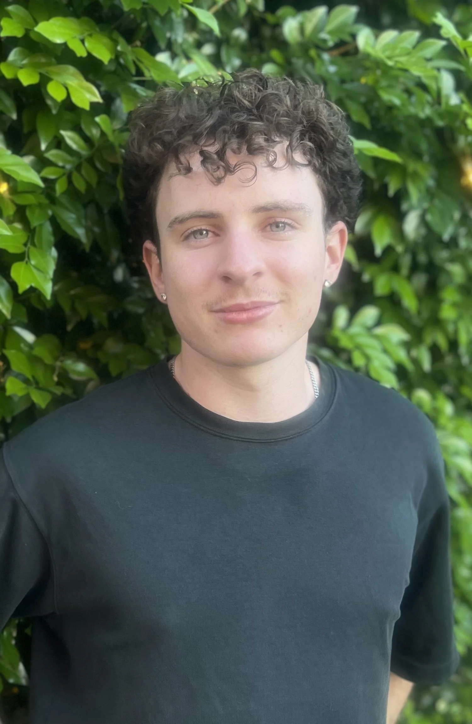 Close-up of a person with short, curly dark hair, light skin, and earrings, standing in front of a green leafy background, wearing a black shirt.