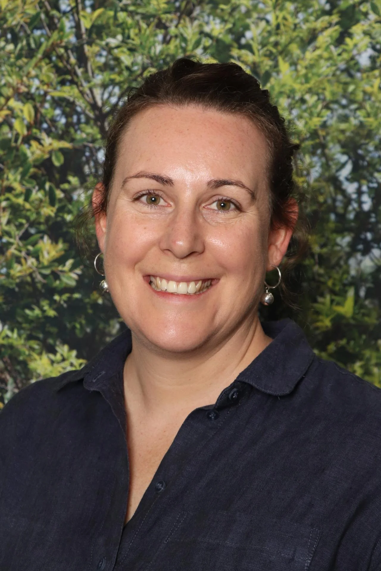 A woman with short dark hair, light skin, green eyes, and earrings smiling in front of a green leafy background.