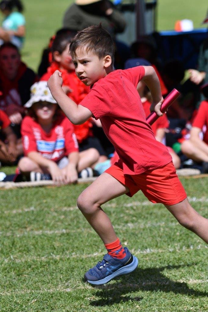 A young boy running on a grassy track during a sports event, holding a relay baton, dressed in a red t-shirt, red shorts, and blue running shoes, with an audience of children and adults in the background.