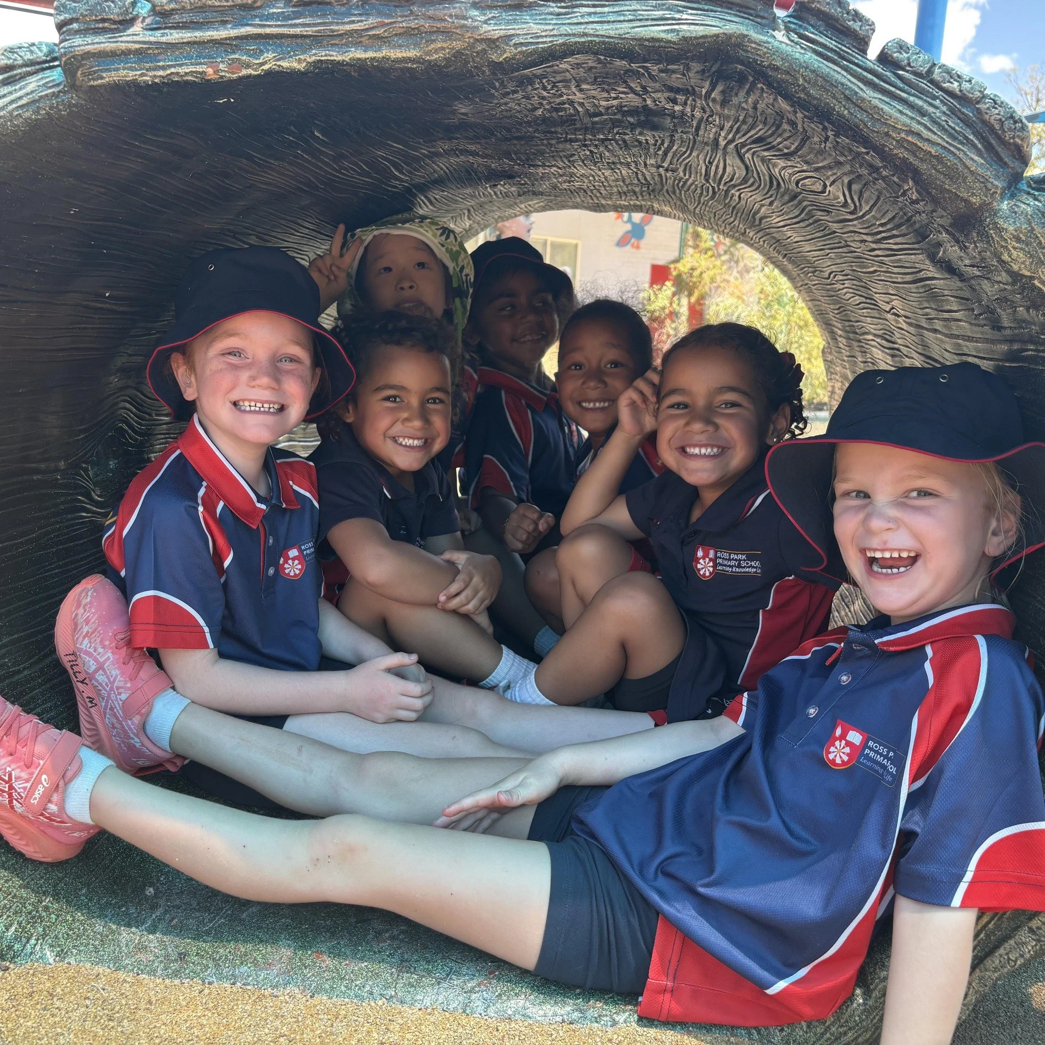A group of smiling children wearing school uniforms and hats, huddled together under a large, textured, metallic or carved overhang at an outdoor playground.