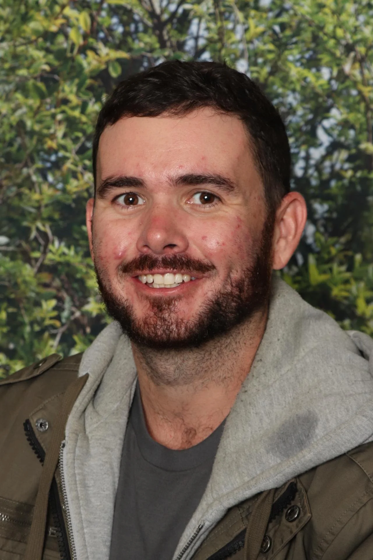A smiling man with dark hair and a beard, wearing a gray hoodie and a jacket, standing outdoors with green foliage in the background.