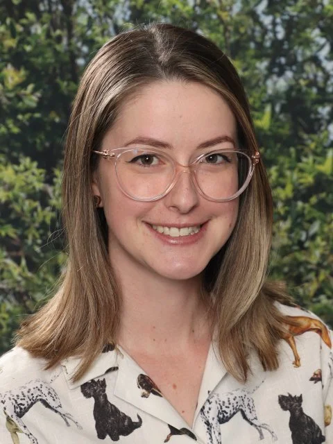 Young woman with glasses smiling outdoors with lush green trees in the background.