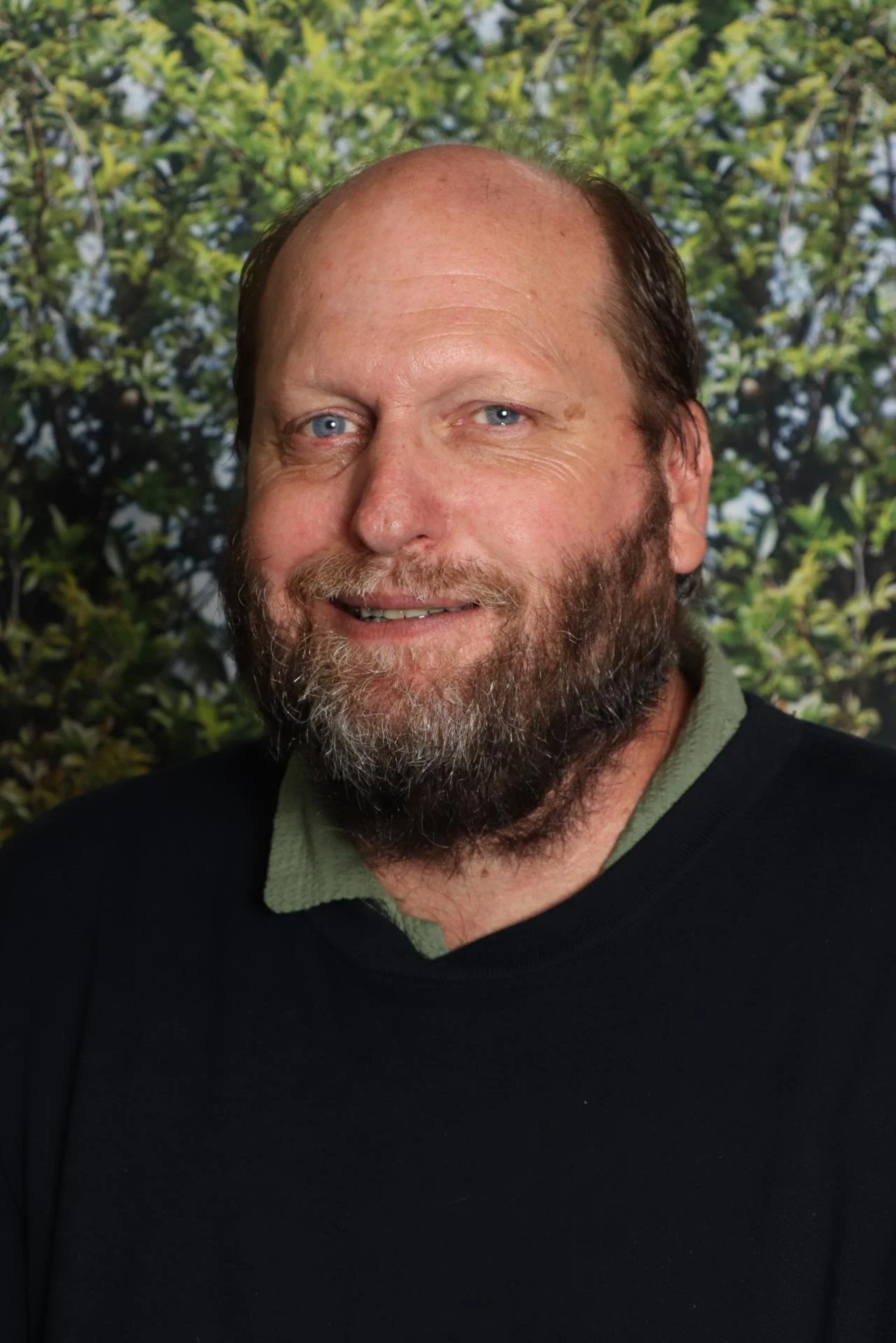 A middle-aged man with a beard and light blue eyes smiling, wearing a black sweater over a green collared shirt, standing outdoors with a blurred background of green foliage.