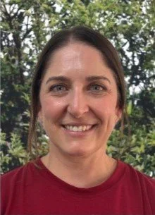 A smiling woman with shoulder-length brown hair, wearing a red shirt, outdoors with green trees in the background.