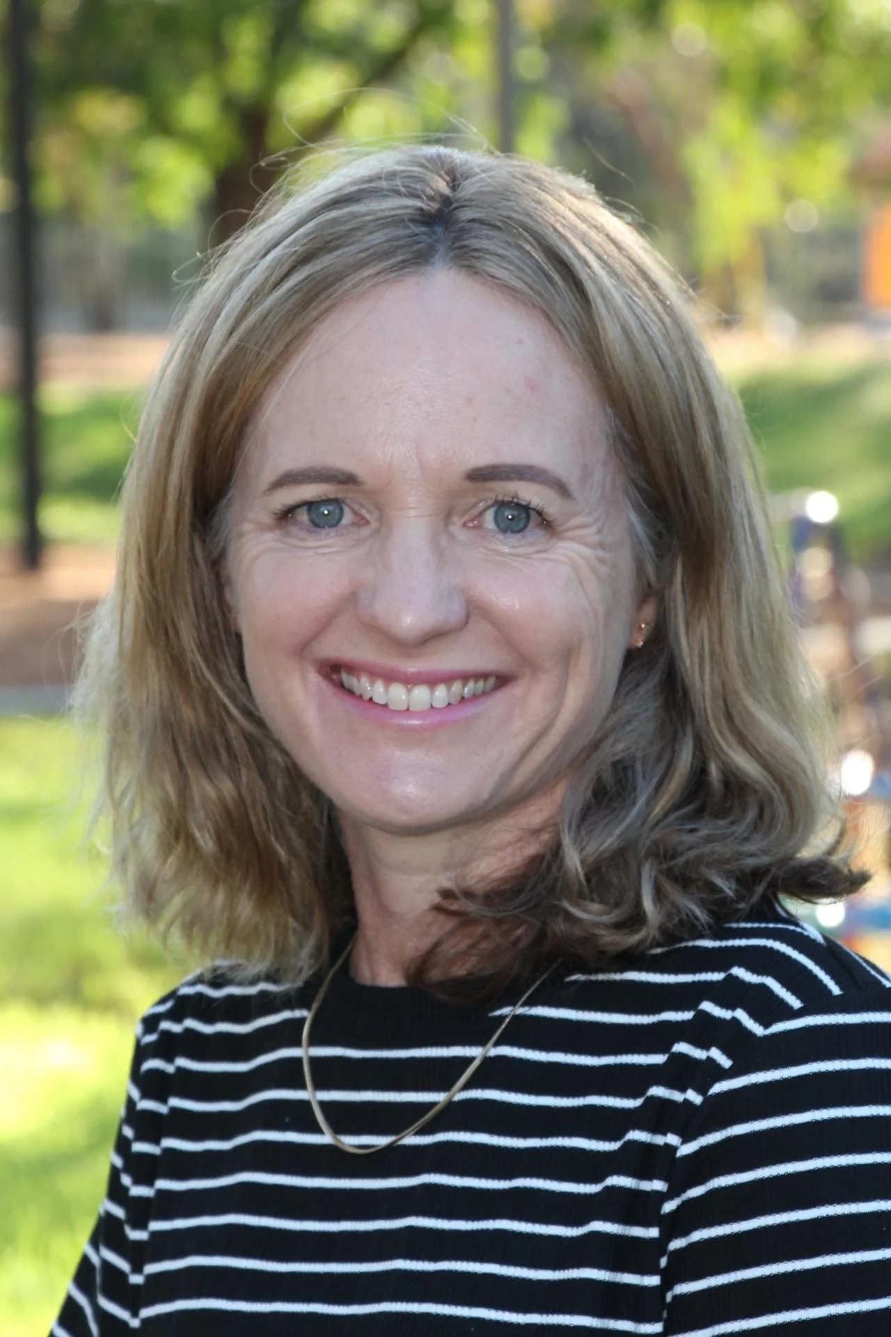A woman with shoulder-length blonde hair, blue eyes, and a big smile, wearing a black and white striped shirt and a gold necklace, outdoors with trees and a park in the background.