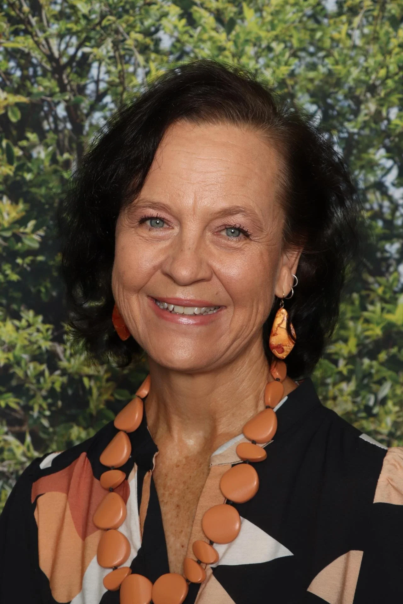 A smiling middle-aged woman with short dark hair and blue eyes, wearing a large peach-colored necklace and matching earrings, standing in front of a leafy green background.