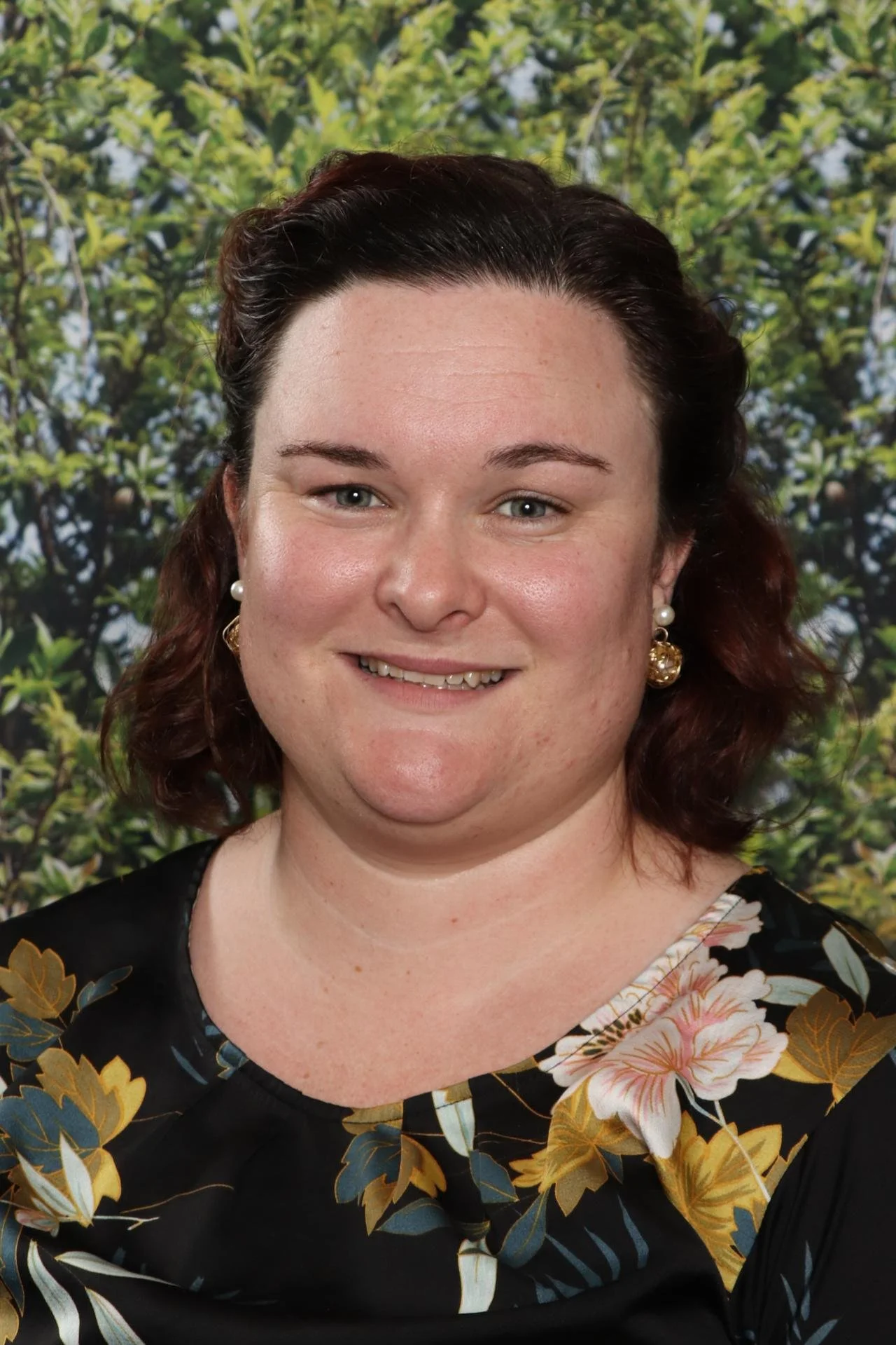 A woman with dark hair, wearing earrings and a black dress with a floral pattern, smiling in front of a leafy green background.