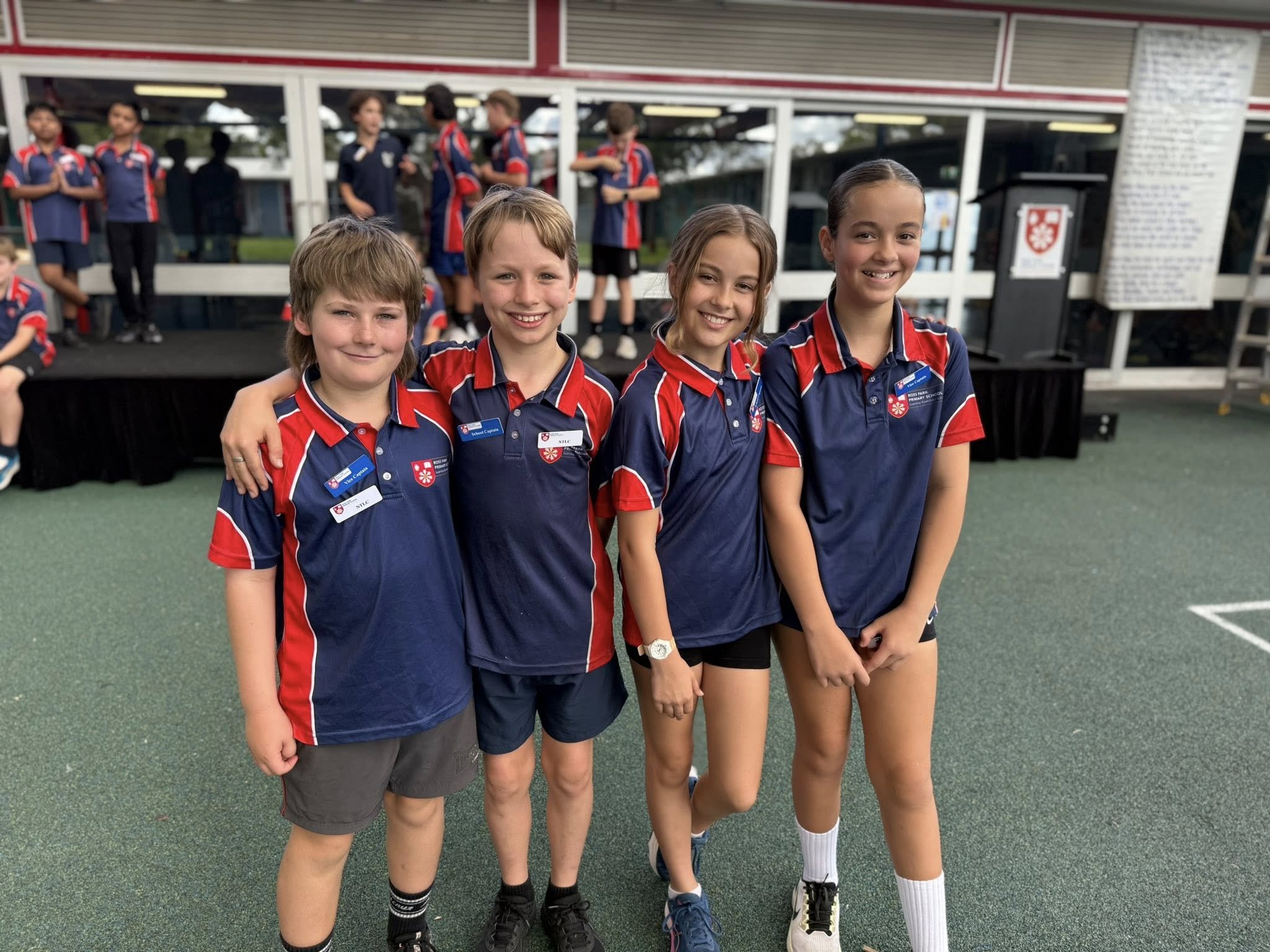 Four children standing together outside on a sports field, wearing uniforms with red, navy, and white colors, smiling at the camera. Four other children are in the background, engaging in conversation near a building with large windows.