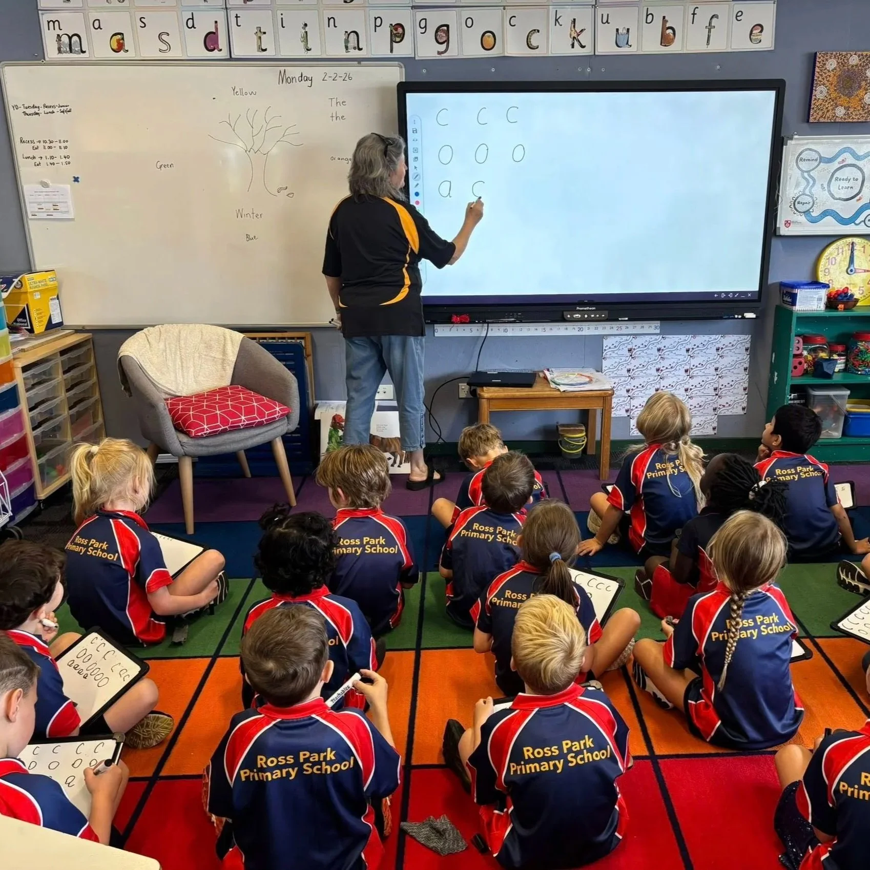 Primary school teacher instructing students on the alphabet and writing on a whiteboard and digital screen, with children sitting on a colorful carpeted floor at Ross Park Primary School.